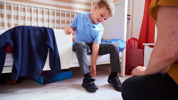 A young smiling blonde boy in a school uniform sitting on his bed and putting on black velcro school shoes