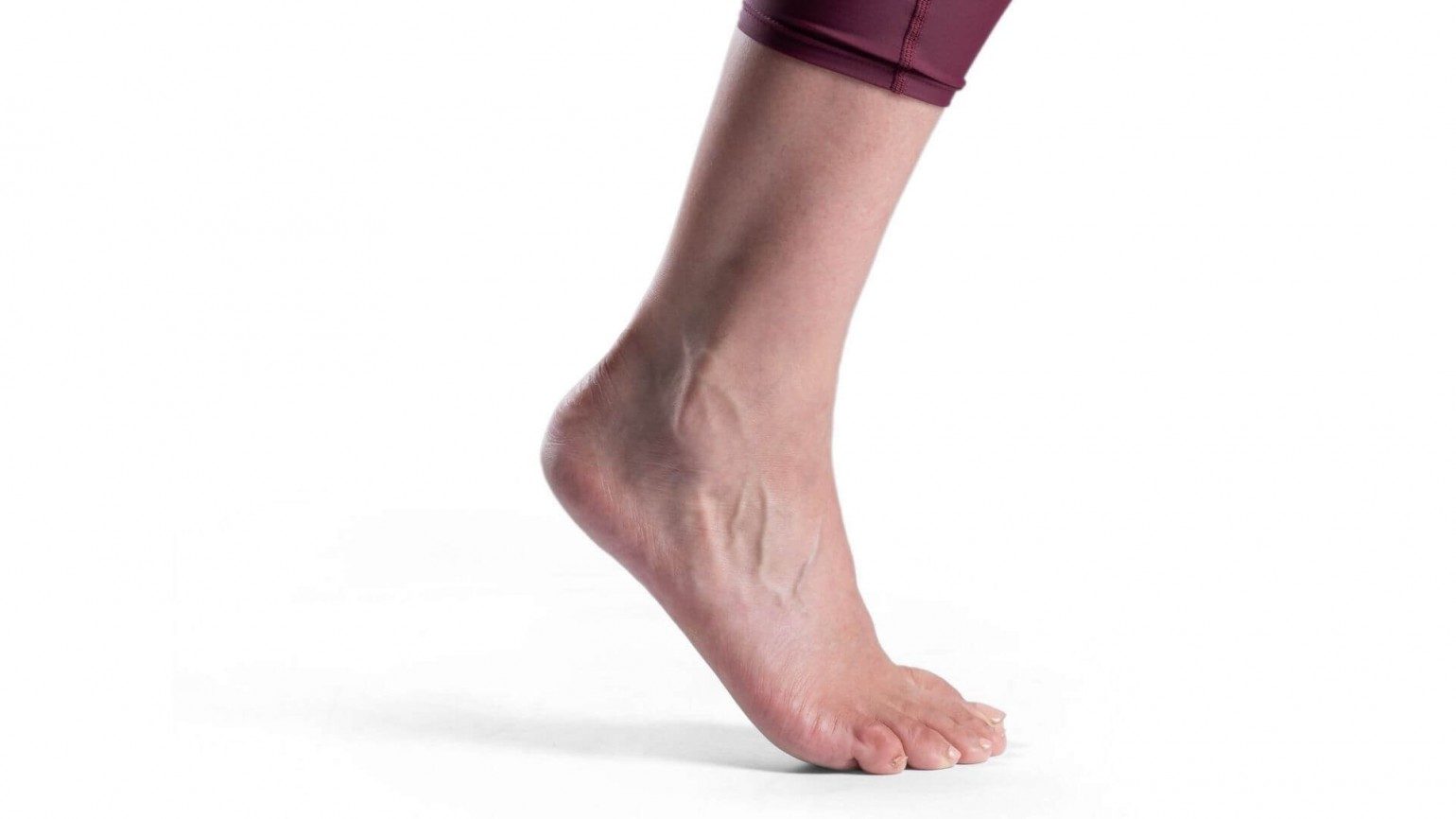 Side view of a white woman's ankle and foot. The ball of her foot is on the ground and the heel is raised in the air. A pair of purple maroon leggings stop just above the ankle