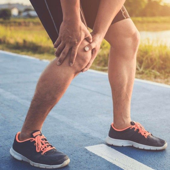 Man in running shoes and exercise shorts standing on a running track clutching his knee in pain