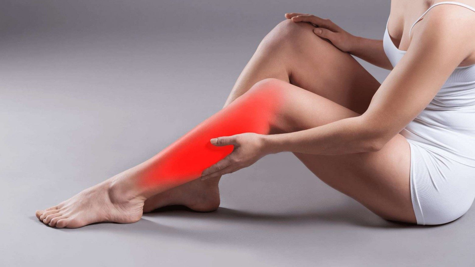 A side view of a woman in white sitting on the floor and touching her lower leg & shin. Her lower leg & shin is highlighted red signifying pain.